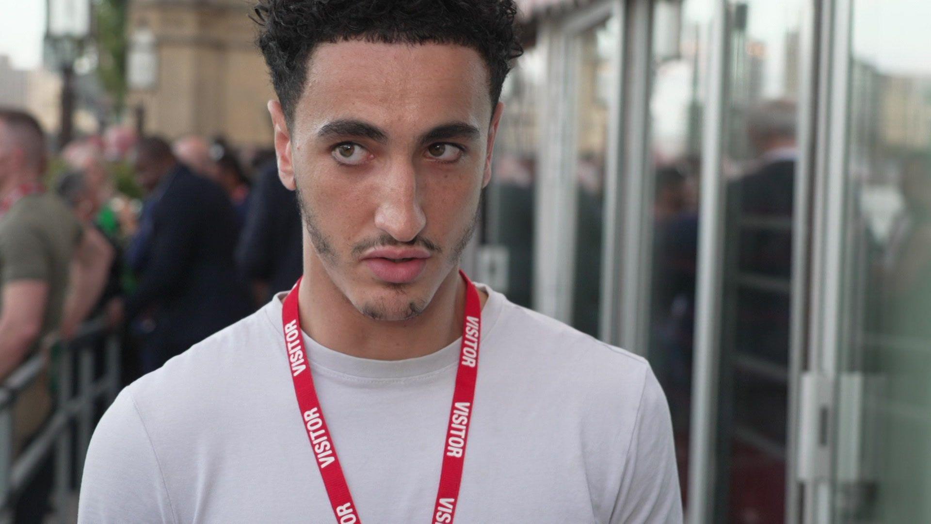 A young man wearing a white T-shirt and a red visitor lanyard stands outside a building, with glass doors and people gathered behind barriers in the background.