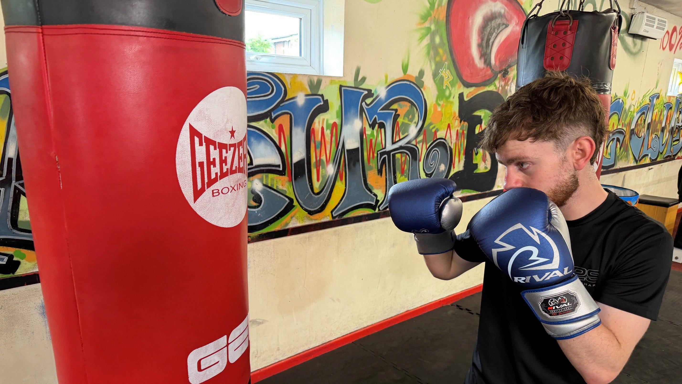 Reuben Muston, wearing a black T-shirt and blue boxing gloves, holds up his firsts and he works out on a red punching bag. He is inside a gym, and the wall behind him has graffiti on it.