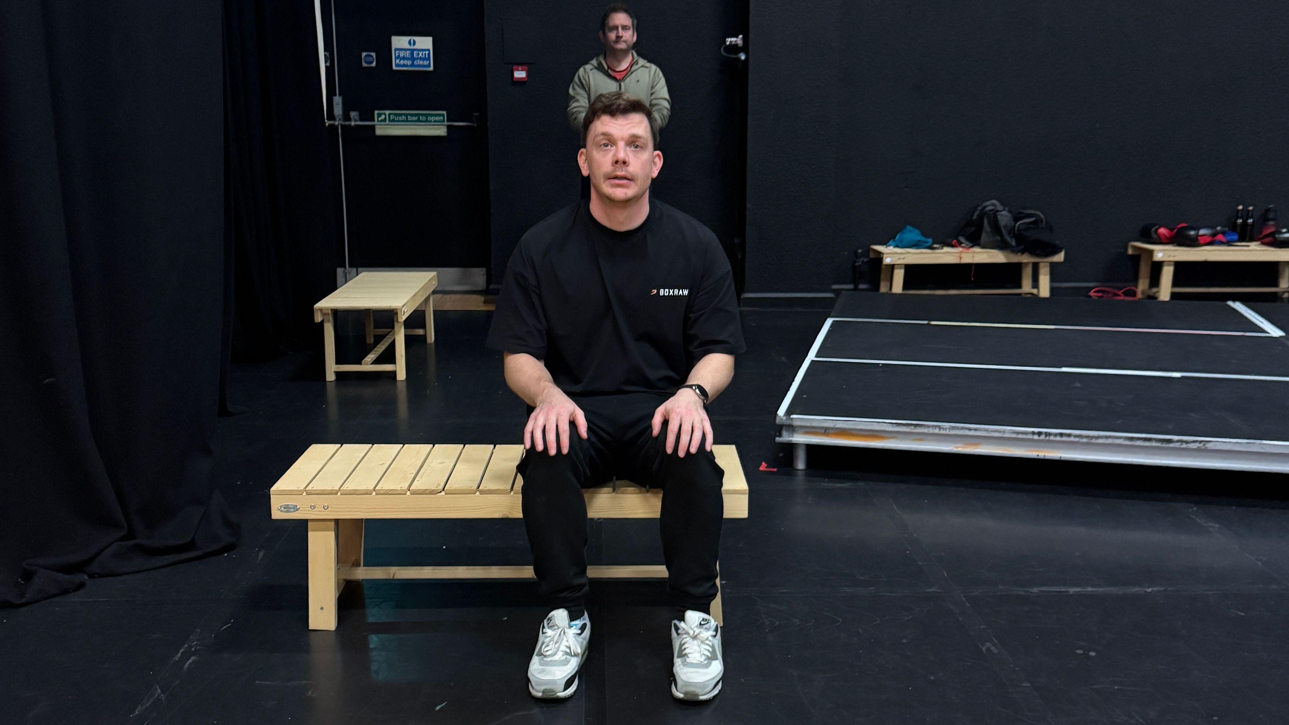 A man sitting on a bench, wearing all black looking above himself to a crowd behind the camera