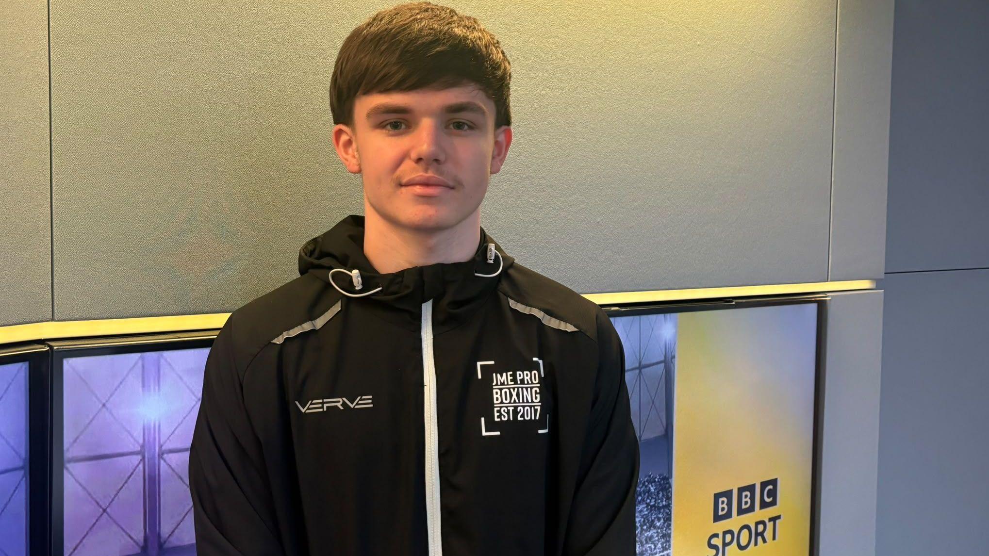 A man with dark hair, wearing a black sports top with JME pro boxing est 2017 printed on the chest. There is a BBC Sport logo to the side of him