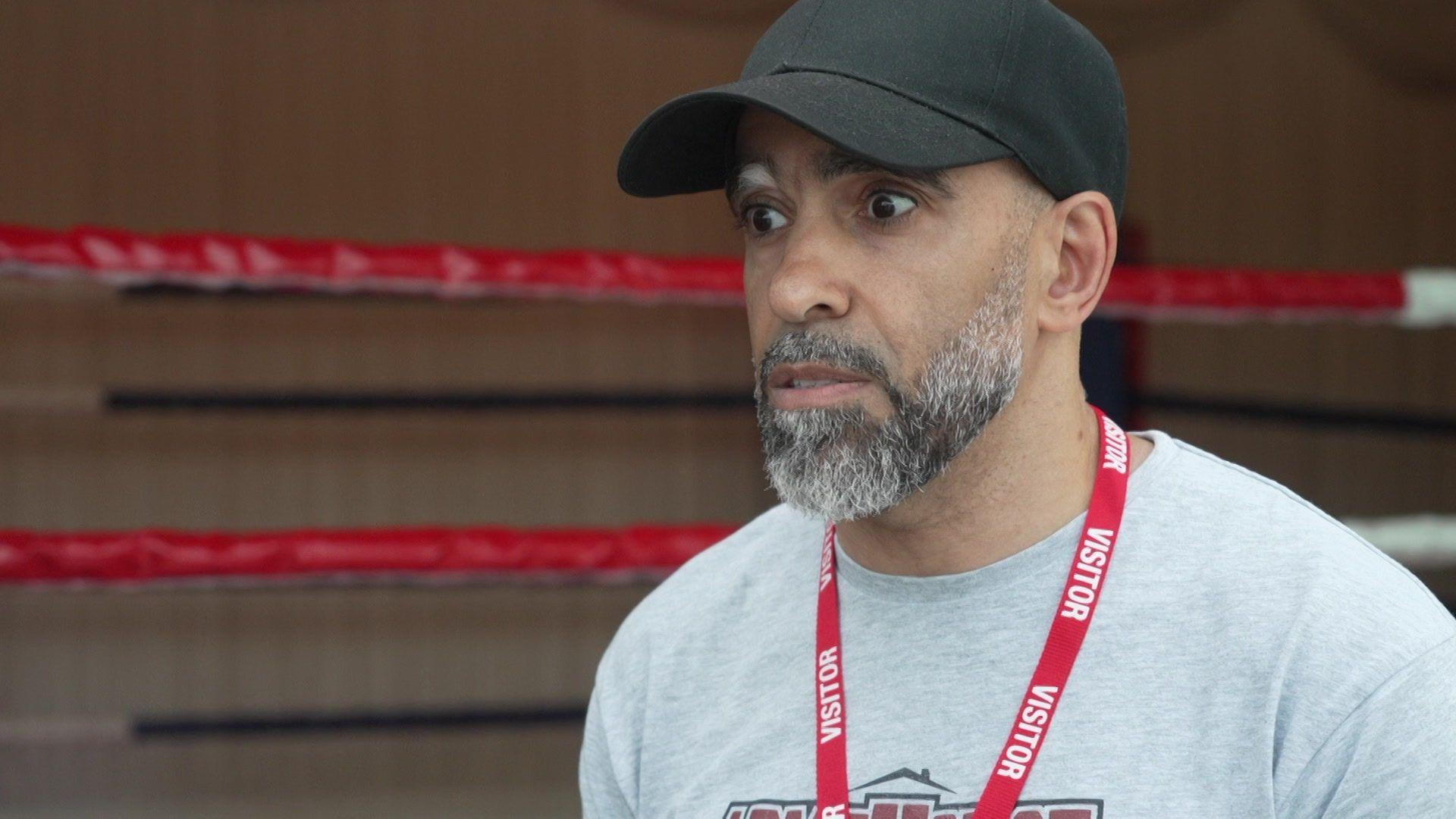 A man wearing a black baseball cap, grey sweatshirt and red visitor lanyard stands beside a boxing ring, with red ring ropes visible in the background.