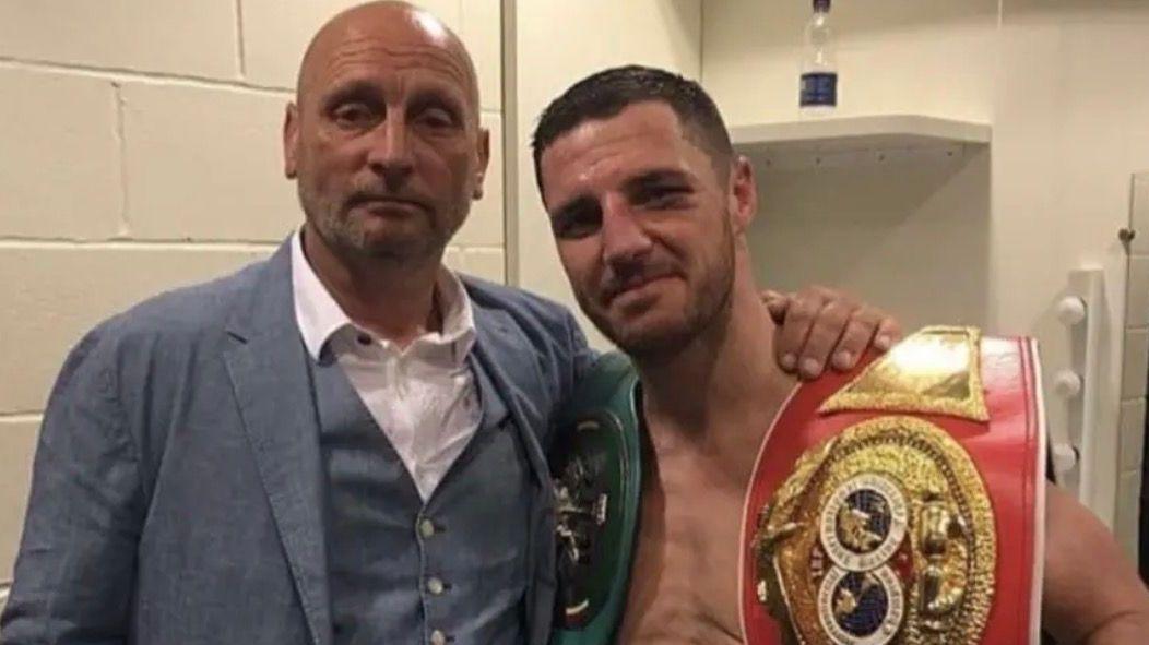 Tommy Coyle is on the right, he's holding a large boxing belt and is shirtless. His eye is bruised as it looks like he's just finished a fight. His dad is on his left in a grey suit with his arm around him. The pair look close.