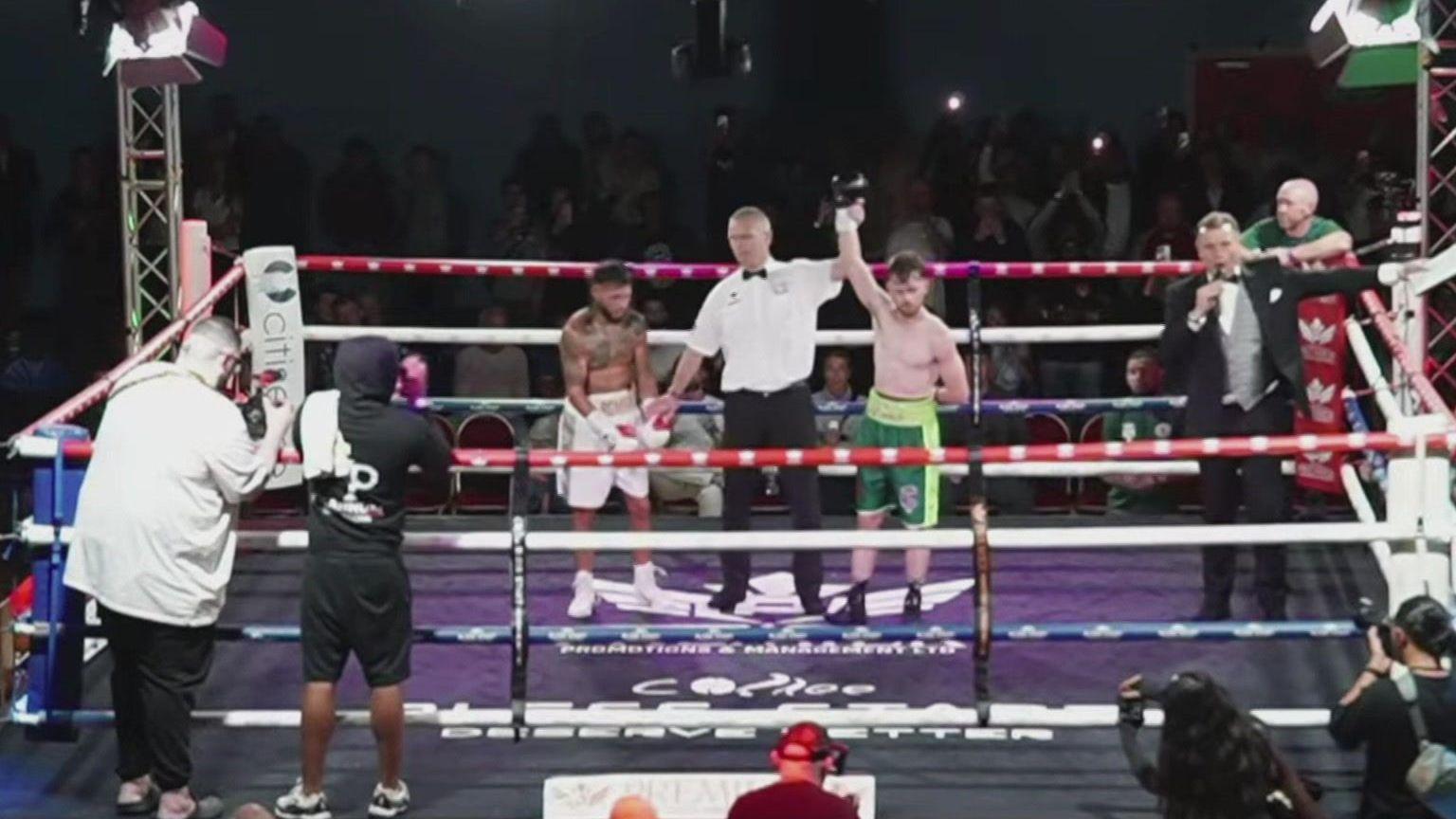 A TV screengrab of a boxing ring with Reuben Muston, in green shorts, having his arm held aloft by the referee