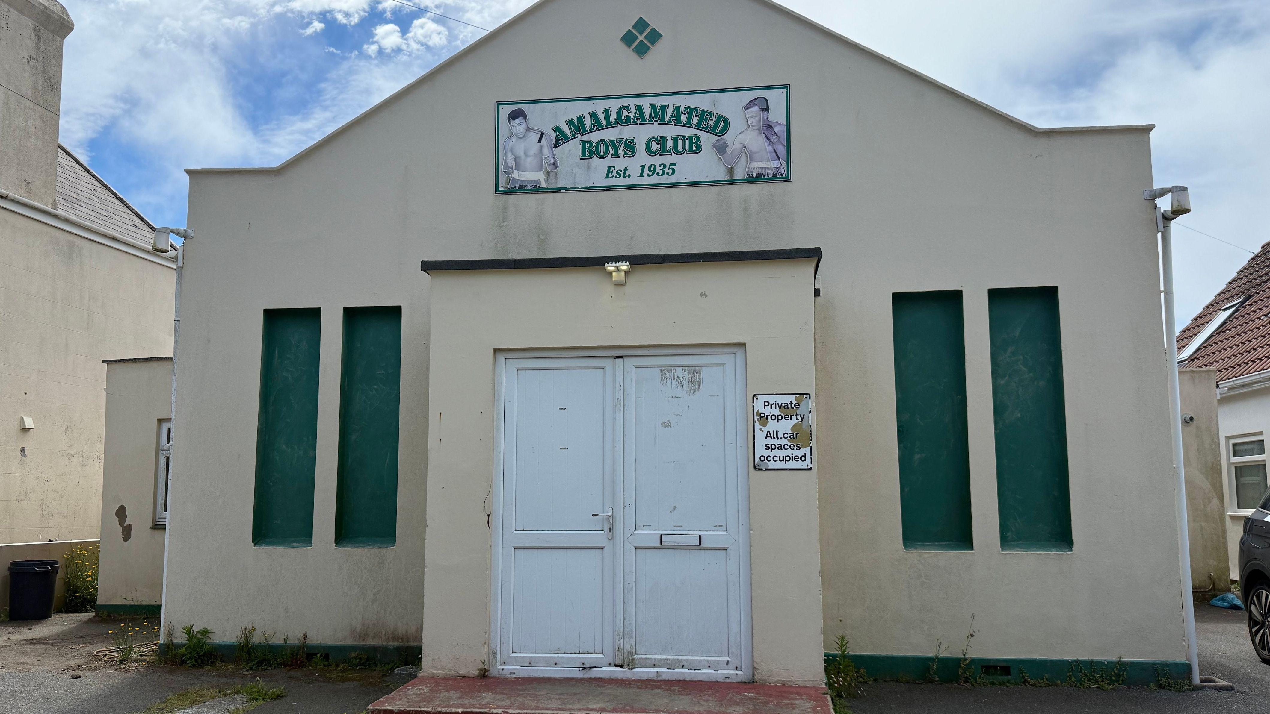 The exterior of the boxing club is a pale white colour. There is a sign for the club above white double doors, and four blocked out windows.