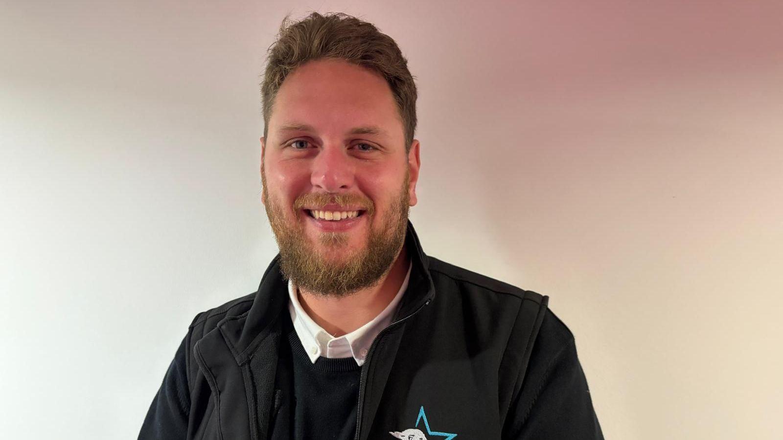 A man with brown hair swept to the side and brown facial hair. He is wearing a black sweater with a white collared shirt underneath, and a black jacket with a blue star on the breast