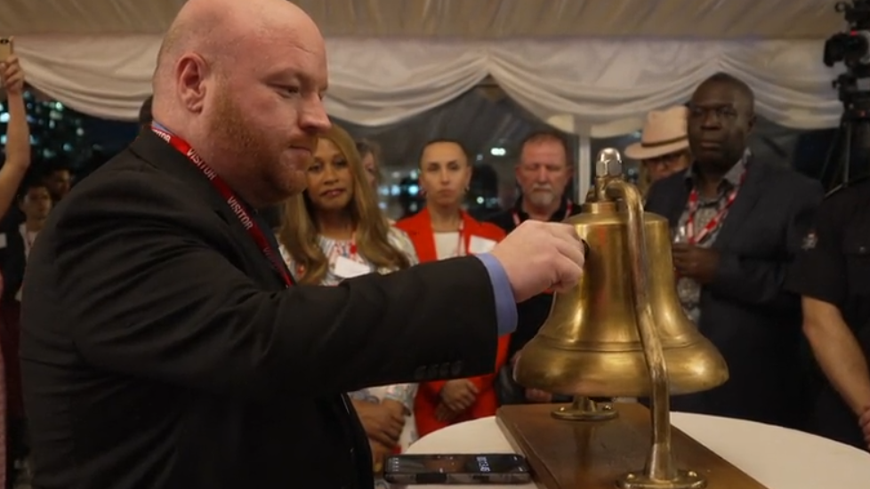 A man in a dark suit rings a large brass bell mounted on a wooden stand while attendees wearing event lanyards gather around inside a marquee-style venue. Several people watch and take photos, with city lights visible in the background.