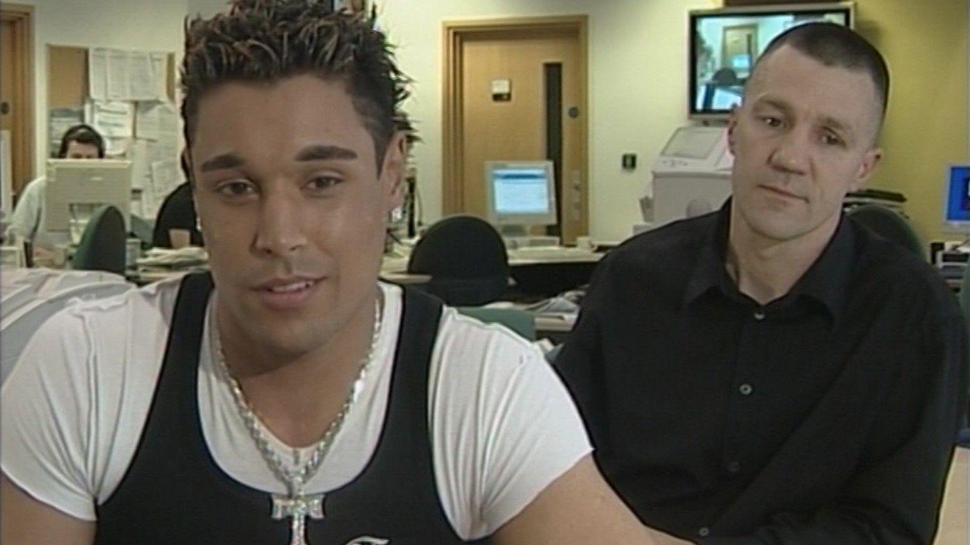 A man with spiky black hair with blonde tints and a silver chain looks into the camera. Another man with short hair wearing a black shirt looks down.