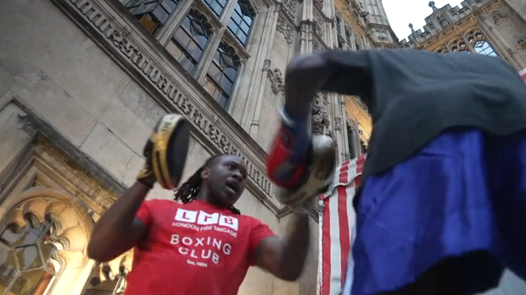 A person wearing a red London Fight Factory boxing club T-shirt holds focus pads while another person throws punches during an outdoor boxing session at Parliament