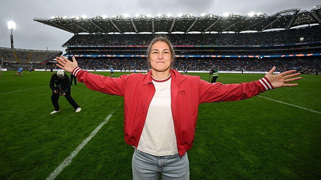 Katie Taylor is wearing a red jacket, white t-shirt and blue jeans and has her arm outstretched as she stands on the pitch at Croke Park