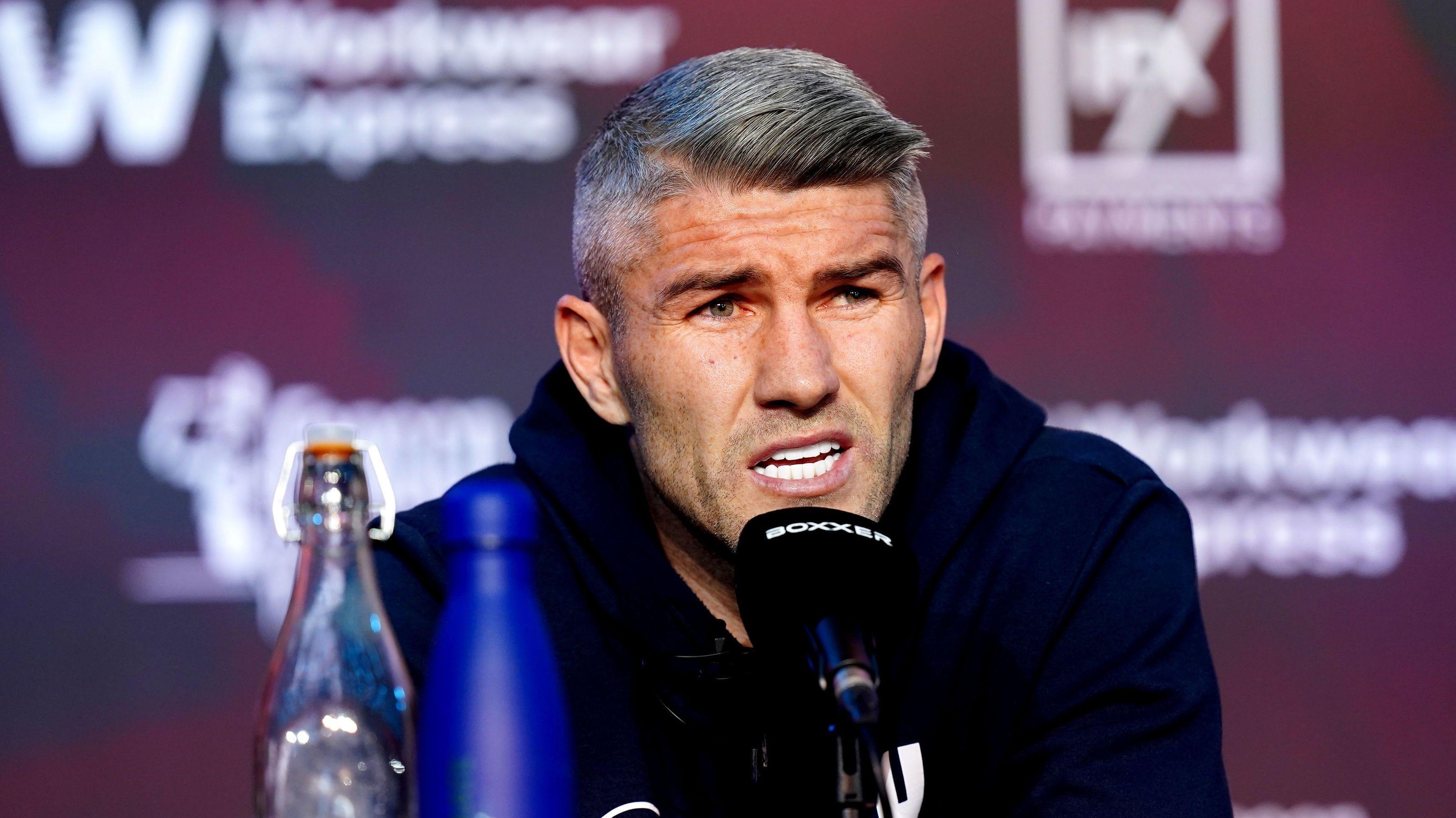 A man in his 30s, with grey hair and wearing a blue hoody, speaks into a black microphone at a press conference.