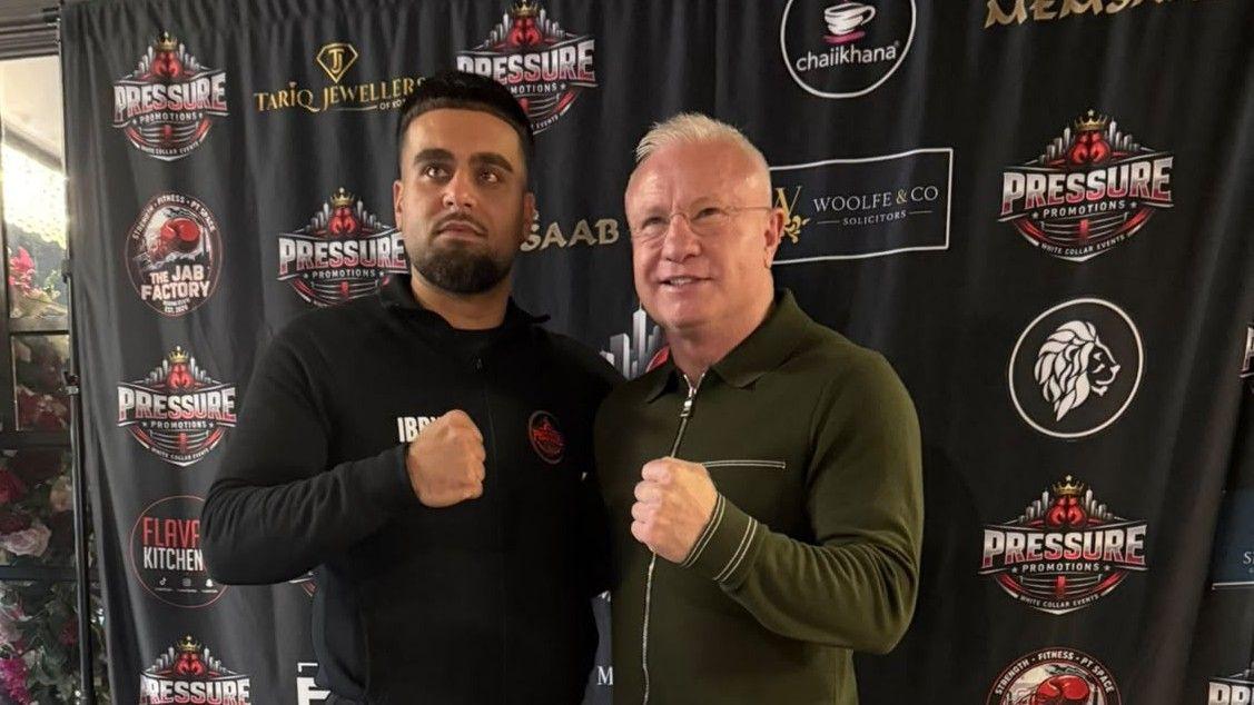 Mehmood and Schwer standing in front of a promotional poster, holding a single fist in front of them in a mock boxing pose. Mehmood has dark hair and a short beard, and is wearing a tight black top.