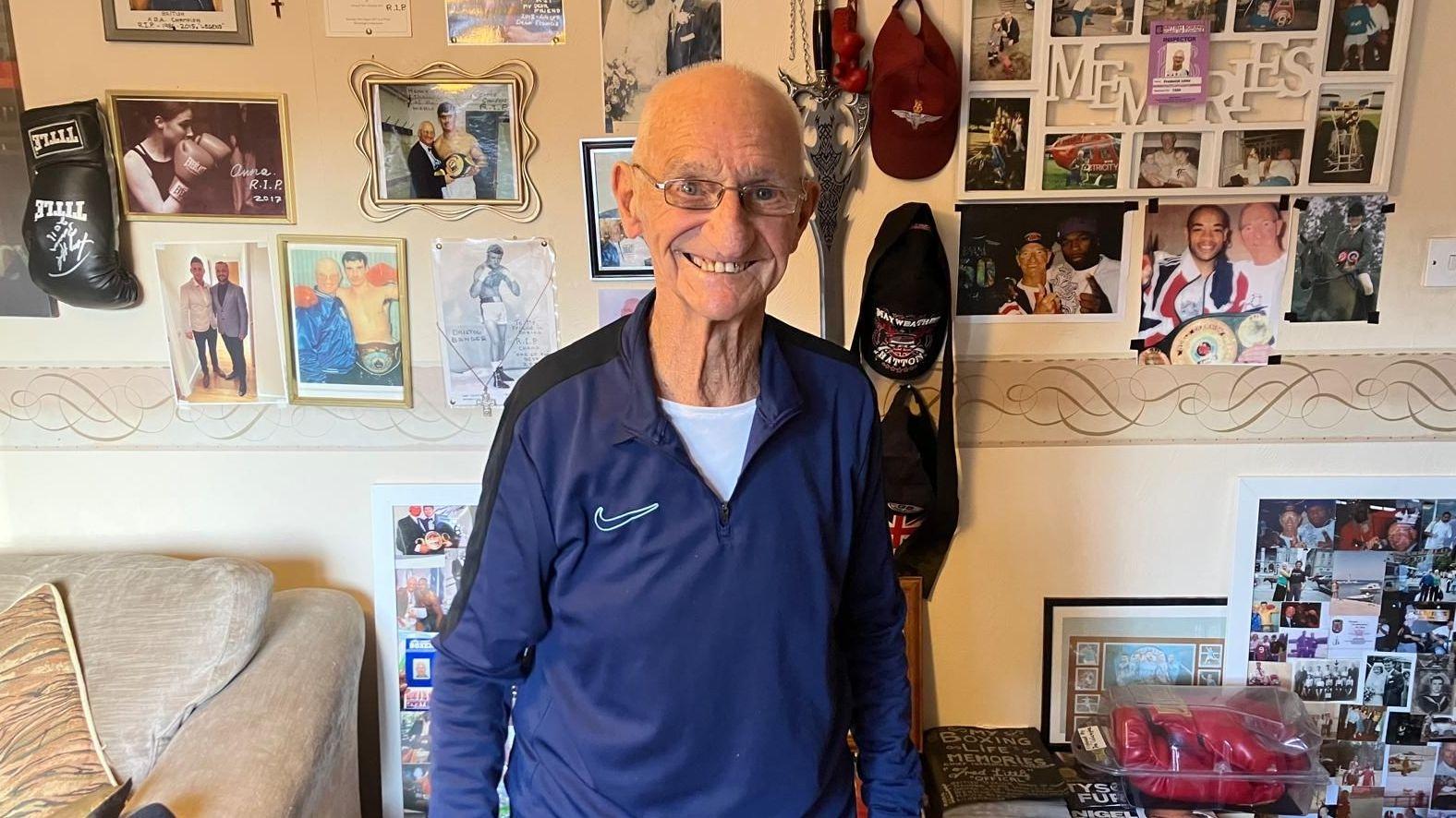 A man in his 90s standing next to a wall full of pictures and artwork from throughout his boxing career.