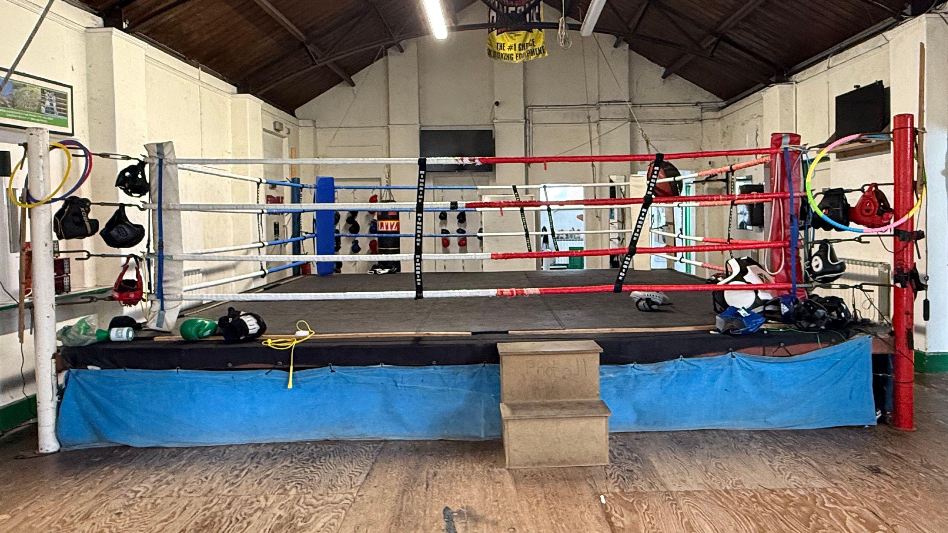 The interior of the boxing club. There is a single boxing ring with a mat and multi-coloured ropes. There are gloves and head protection gear hanging on the ropes.