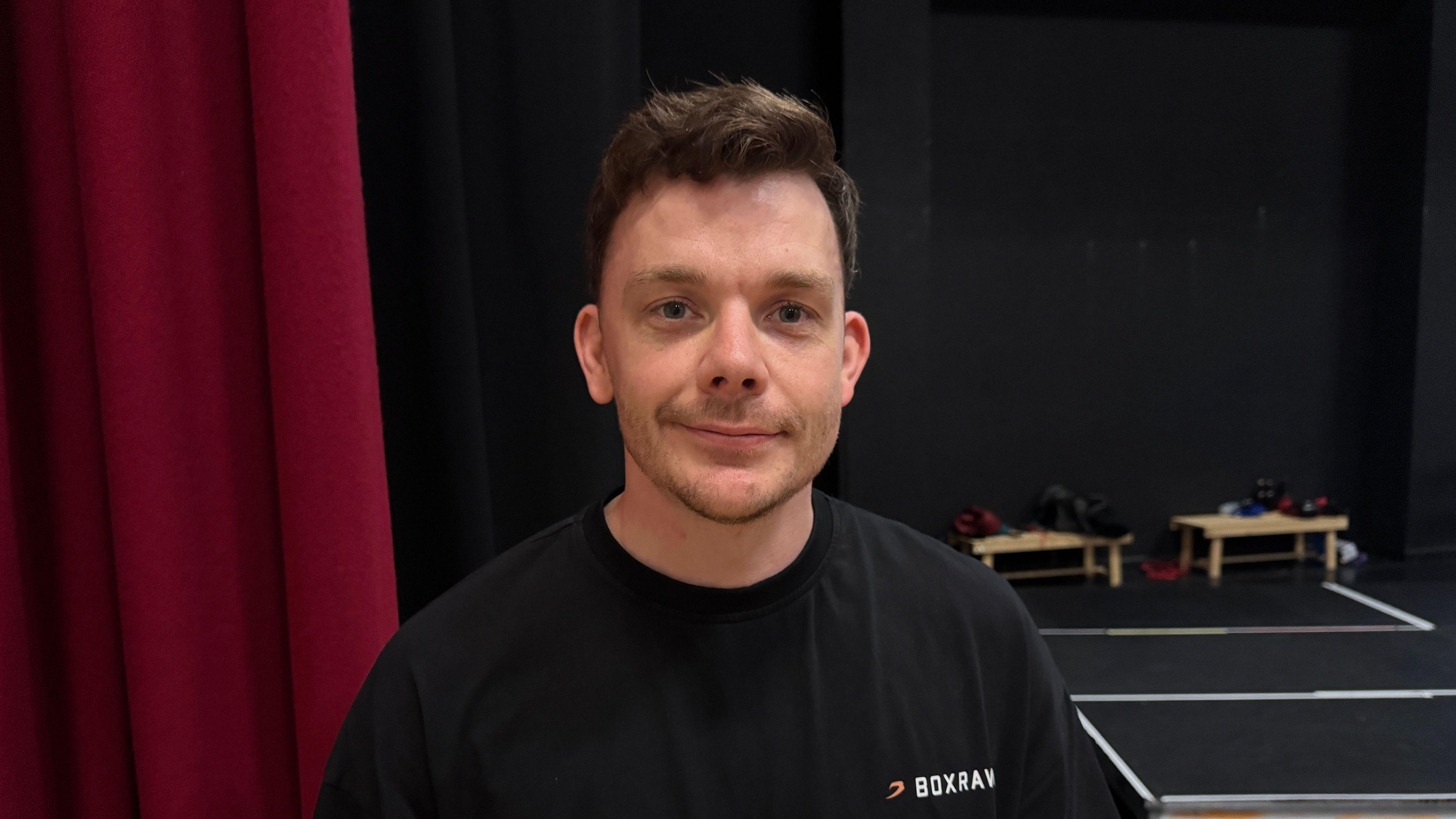 A man looking straight at the camera smilling - he is wearing a black t-shirt.